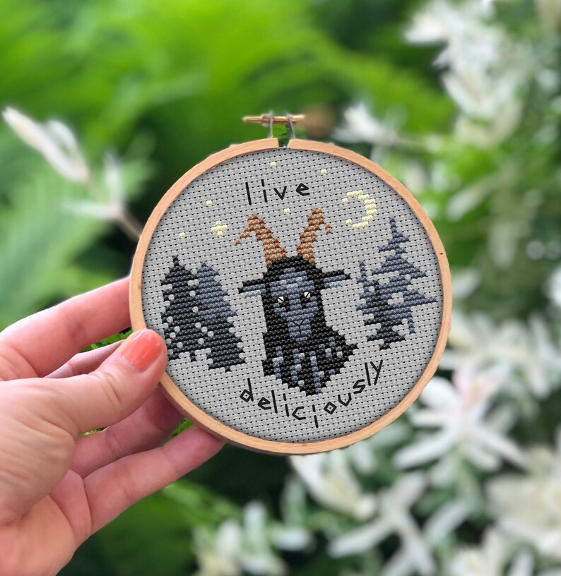 May include: A grey and black embroidery hoop with a black goat with horns and the text "Live deliciously" in black.