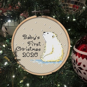 May include: A circular wooden embroidery hoop containing a cross-stitch design of a white polar bear sitting on blue ice. The text reads Baby's First Christmas 2026 in black lettering, with small white squares representing falling snow.