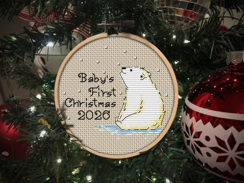 May include: A circular wooden embroidery hoop containing a cross-stitch design of a white polar bear sitting on blue ice. The text reads Baby's First Christmas 2026 in black lettering, with small white squares representing falling snow.