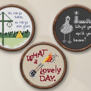 May include: Three framed cross-stitch embroidery hoops with different designs. The first hoop features a green and yellow camping scene with the text "As Härga takes, so Härga also gives". The second hoop features a gray bird with a lighthouse and the text "Why'd ye spill yer beans?". The third hoop features a red and orange fire with the text "What a lovely DAY."
