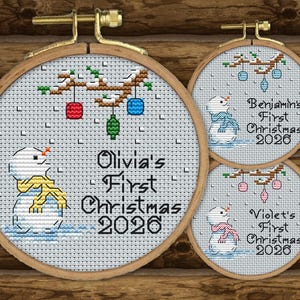 May include: Three circular wooden embroidery hoops containing cross-stitch designs of snowmen with colorful scarves and hanging ornaments. The text reads Olivia's First Christmas 2026, Benjamin's First Christmas 2026, and Violet's First Christmas 2026.