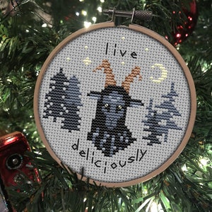 May include: A grey and black cross-stitch ornament featuring a goat in the centre, surrounded by trees and a crescent moon. The text "I live deliciously" is stitched below the goat.