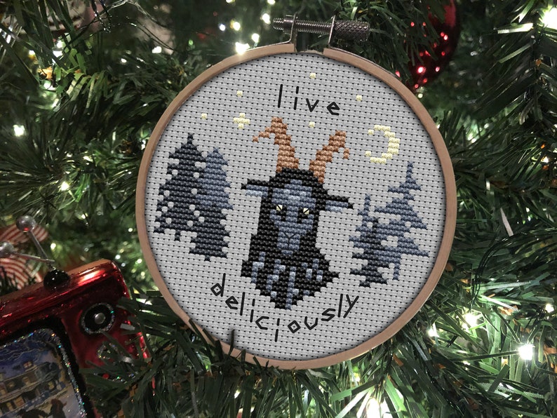 May include: A grey and black cross-stitch ornament featuring a goat in the centre, surrounded by trees and a crescent moon. The text "I live deliciously" is stitched below the goat.