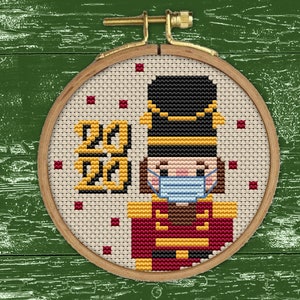 May include: A cross-stitch design of a nutcracker wearing a blue face mask with the year 2020 stitched in yellow thread.