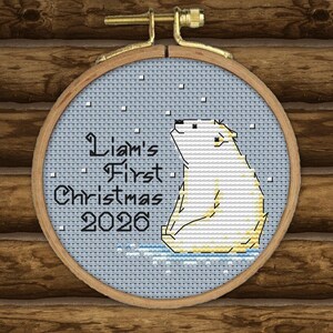 May include: A circular wooden embroidery hoop containing a cross-stitch design of a white polar bear sitting on a light blue background with falling snow. The text reads Liam's First Christmas 2026 in black lettering on the left side of the bear.