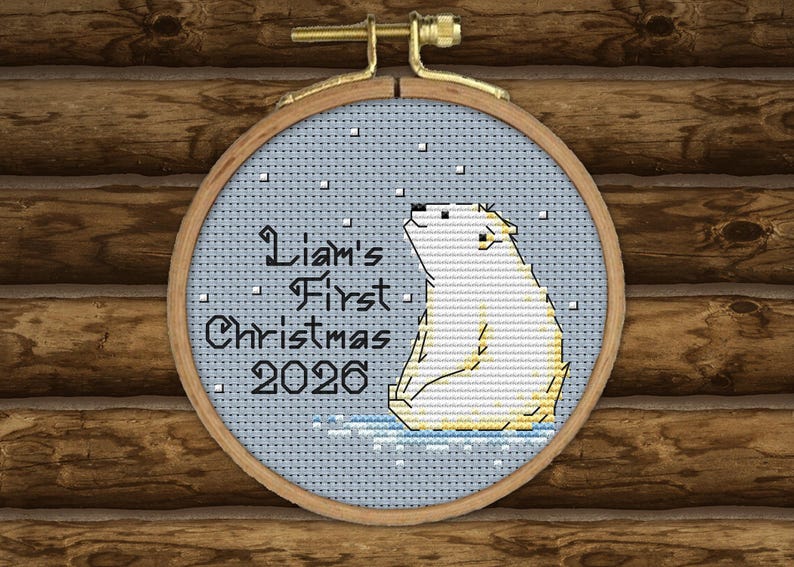 May include: A circular wooden embroidery hoop containing a cross-stitch design of a white polar bear sitting on a light blue background with falling snow. The text reads Liam's First Christmas 2026 in black lettering on the left side of the bear.