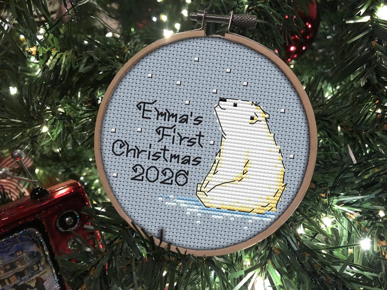 May include: A circular wooden embroidery hoop containing a cross-stitch design of a white polar bear sitting on blue ice. The text reads Emma's First Christmas 2026 in black lettering against a light blue background with small white squares.