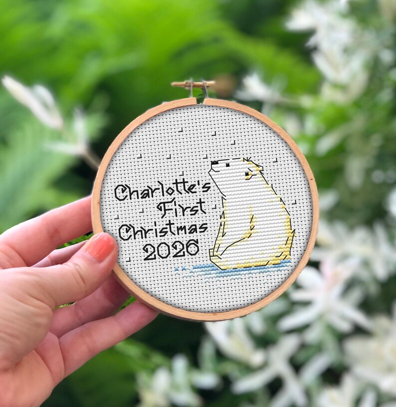 May include: A circular wooden embroidery hoop containing a cross-stitch design on white fabric. The design features a yellow-outlined polar bear sitting on a blue patch, with the black text "Charlotte's First Christmas 2026" printed to the left.