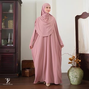 May include: A dusty rose-coloured abaya featuring a loose, flowing silhouette with long sleeves and an integrated matching hijab. The garment is made of a solid, smooth fabric and reaches the floor, providing full coverage with a simple design.
