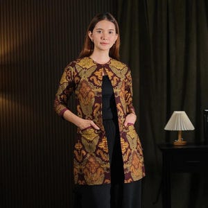 Peut inclure: Une veste ouverte à manches longues avec un motif batik marron foncé et doré, ornée de détails floraux et de papillons. Le vêtement présente deux poches plaquées à l'avant avec un liseré rouge foncé et une encolure ronde.