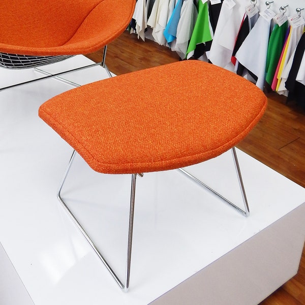 May include: Orange upholstered ottoman with a chrome wire frame. The ottoman is sitting on a white surface.