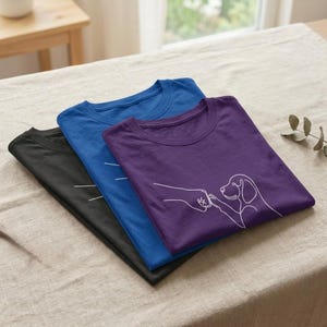 Può includere: Tre magliette piegate in nero, blu e viola. Ogni maglietta presenta una grafica a linee bianche che raffigura una mano umana che fa un pugno con una zampa di cane. Le magliette sono impilate, mostrando il design a linea continua.
