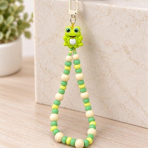Può includere: Un ciondolo per telefono con una perlina a forma di rana verde attaccata a un anello di perline sferiche alternate color crema, verde lime e verde bosco. L'accessorio include un moschettone in plastica bianca per il fissaggio al dispositivo.