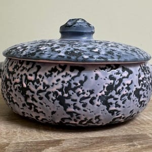 May include: A round ceramic casserole dish with a matching lid, featuring a textured, mottled glaze in shades of black, light purple, and soft pink. The pot has two small side handles and a central knob on the lid, showcasing a speckled finish.