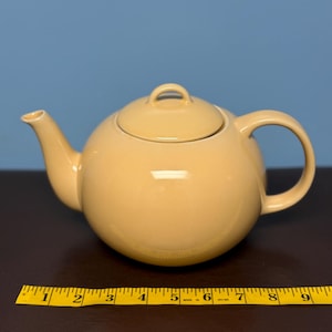 May include: A round, pale yellow ceramic teapot with a matching lid, a curved spout, and a handle. A yellow measuring tape is positioned in front of the teapot, showing a width of approximately 23 cm from the spout to the handle edge.