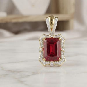 May include: A rectangular pendant featuring a large, deep red gemstone set in yellow gold. The frame and the bail are decorated with small, clear pavé-set stones. The pendant has a distinct, ornate shape with pointed corners and a polished gold finish.