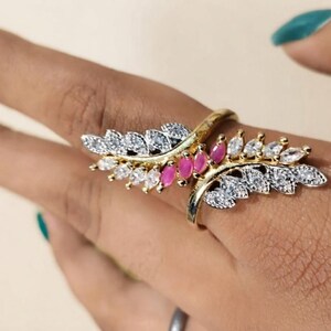 May include: A gold-toned ring featuring two overlapping leaf-shaped branches. The design is embellished with marquise-cut clear white stones and a central row of marquise-cut pink stones, creating a symmetrical, nature-inspired decorative piece.