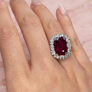 May include: A ring featuring a large, rectangular, deep red gemstone set in a silver-toned metal band. The central red stone is surrounded by a halo of numerous small, clear, sparkling white stones arranged in a dense, oval-shaped cluster.