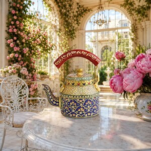 May include: A decorative metal tea kettle featuring a vibrant hand-painted floral pattern in shades of deep blue, gold, and red. The kettle has a curved red handle with gold detailing, a matching lid, and a spout, all adorned with intricate designs.