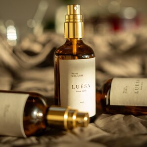 Puede incluir: Tres botellas de vidrio ámbar con boquillas rociadoras doradas y etiquetas blancas. La botella central presenta el texto No. 03 MILANO LUESA Room Spray. Las botellas están dispuestas en grupo, una de pie y dos acostadas horizontalmente.