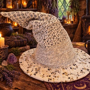 May include: A quilted witch hat featuring a white fabric base with a black floral and paisley print. The hat has a wide, circular brim and a tall, pointed, slightly slumped crown, constructed with visible stitching throughout the fabric.