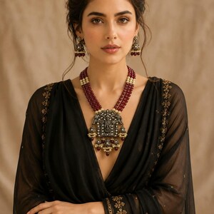 May include: A multi-strand necklace featuring deep red beads and a large, ornate, oxidized silver-toned pendant with intricate engravings and small dangling beads. Matching bell-shaped jhumka earrings with red and white accents complete the set.