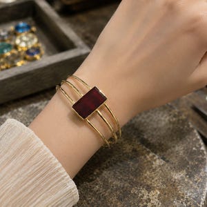 Può includere: Un bracciale rigido color oro caratterizzato da tre sottili fasce parallele e un inserto centrale rettangolare in pietra rosso scuro. Le fasce metalliche sono lisce e curvate per adattarsi al polso, offrendo un aspetto strutturato.