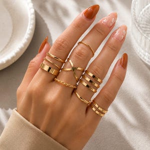 Op de afbeelding: Een set goudkleurige ringen gedragen aan meerdere vingers. De collectie bevat dunne gladde banden, een gedraaide touw-stijl band, een brede platte band, een drielaagse band, een dubbellaagse band en een ring met een centraal kruisontwerp.
