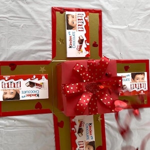 May include: A red square gift box with a red ribbon featuring gold hearts, surrounded by four open flaps. Each flap displays a Kinder Chocolate bar box with the text Kinder Chocolate and a child's face, decorated with red hearts and gold stars.