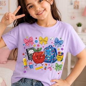 May include: A light purple t-shirt featuring a colorful graphic print of school supplies including a red apple, a blue backpack, a notebook, a pencil, crayons, scissors, glue, and various bows, all designed with smiling faces and bright patterns.