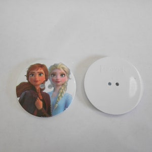 May include: Two white round buttons with a printed image of the characters Anna and Elsa from the movie Frozen. One button has the text "Kamica" printed on it.