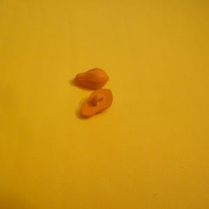 Puede incluir: Dos pequeños botones de arcilla hechos a mano, de color naranja, sobre un fondo amarillo. Un botón está entero, mientras que el otro está cortado por la mitad, revelando un agujero en el centro. Los botones tienen un acabado liso y mate.