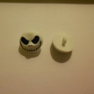 Könnte beinhalten: Zwei weiße Plastikknöpfe mit einem schwarzen Jack Skellington Gesichtsdesign. Ein Knopf hat eine Schlaufe auf der Rückseite zum Nähen.