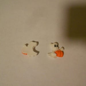 May include: Two white ghost figurines, one holding a small orange pumpkin. The ghost on the left is a simple outline, while the ghost on the right has a face and is holding the pumpkin in its arms.