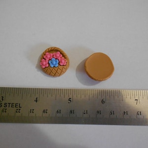 May include: Two small, round, brown polymer clay embellishments. One is a basket with pink and blue flowers, the other is a plain circle. The basket is approximately 1 inch in diameter.