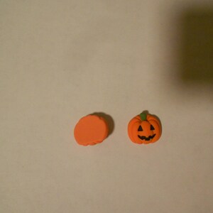 May include: Two orange Halloween decorations. One is a small, smiling jack-o'-lantern with a green stalk. The other is a flat, orange, circular shape. Both are on a plain, off-white background.