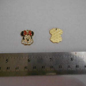 Op de afbeelding: Twee goudkleurige bedels. Eén bedel is een Minnie Mouse-hoofd met een rode strik, zwarte oren en een crèmekleurig gezicht. De andere bedel is een goudkleurige shirtvorm met een textuurpatroon. Een liniaal staat in de afbeelding.