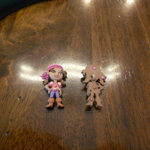 Può includere: Due piccole figurine di plastica di una pirata. Una figurina è a colori con una bandana rosa, una camicia viola e stivali marroni. L'altra figurina è marrone con accenti rosa.