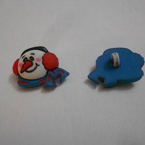 May include: A pair of plastic buttons featuring a snowman with a red scarf and earmuffs. The back of the button is blue.