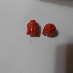 May include: Two red ceramic beads with a Buddha face design. The beads are smooth and have a glossy finish.