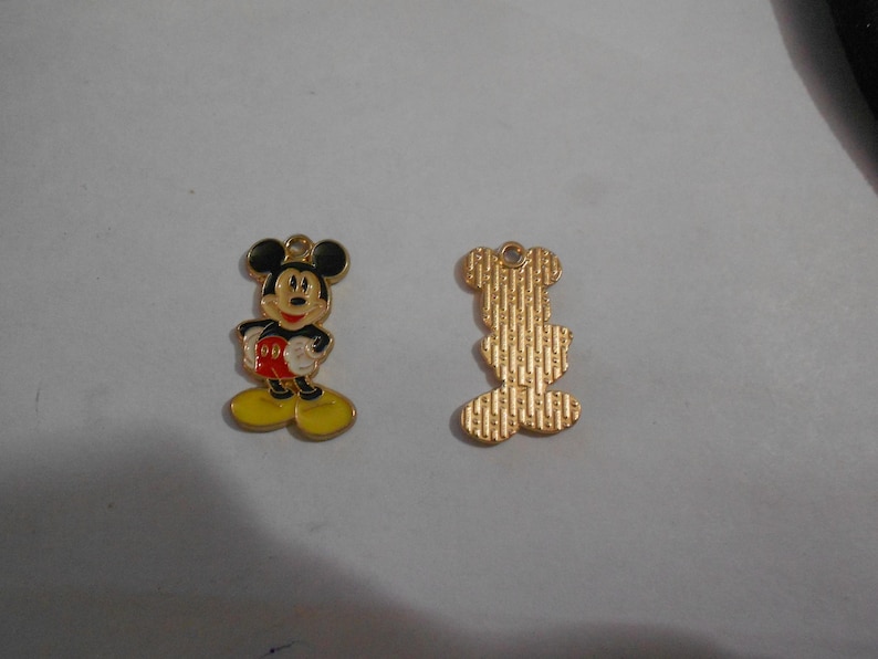 Puede incluir: Dos colgantes dorados de Mickey Mouse. Un colgante est&aacute; completamente coloreado con orejas negras, una camisa roja, zapatos amarillos y una cara sonriente. El otro colgante es la parte trasera de Mickey Mouse, con un acabado dorado texturizado.