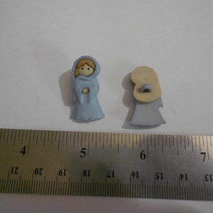 May include: Two small, light blue and beige buttons in the shape of a person wearing a hooded robe. The buttons are next to a metal ruler with inch measurements.
