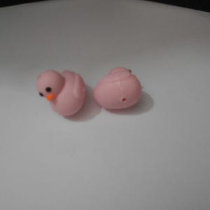 May include: Two small, pink duck figurines with black eyes and orange beaks. One duck is upright, while the other is lying on its side. The ducks are on a white surface.