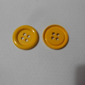 Pode incluir: Dois botões amarelo brilhante, cada um com quatro furos. Os botões são redondos com uma borda ligeiramente elevada. São feitos de um material liso e brilhante sobre um fundo branco.