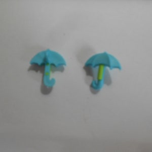 May include: Two small, light blue umbrella-shaped charms. The handles are a lighter blue with a curved hook shape. One umbrella has a green handle, the other has a yellow-green handle. The umbrella tops have a ridged design.