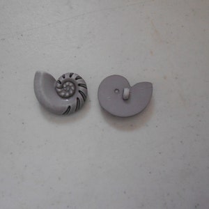 May include: A pair of gray shell-shaped buttons with a black outline. One button has a detailed spiral design, while the other is plain.