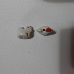 May include: Two small, white ceramic bird figurines. One is a stylized chicken shape, the other a bird head. Both have red, yellow, and black accents. The chicken has a hole for stringing.
