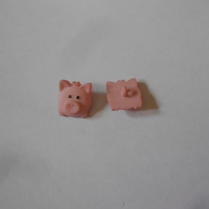 Pode incluir: Dois botões de plástico rosa em forma de porco. Um botão mostra a frente do porco com um focinho e olhos. O outro botão mostra as costas do porco.