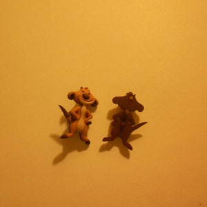 May include: Two small, cartoon meerkat figurines. One is tan and cream-colored with a smiling face and the other is brown. Both have tails and are posed with their hands on their hips. The background is a solid, light yellow.