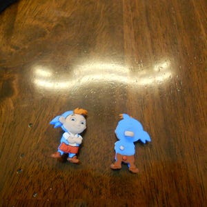 Può includere: Due piccole figurine di personaggi dei cartoni animati blu e arancioni. I personaggi indossano cappelli blu e hanno vestiti rossi e arancioni. Le figurine sono su una superficie di legno.